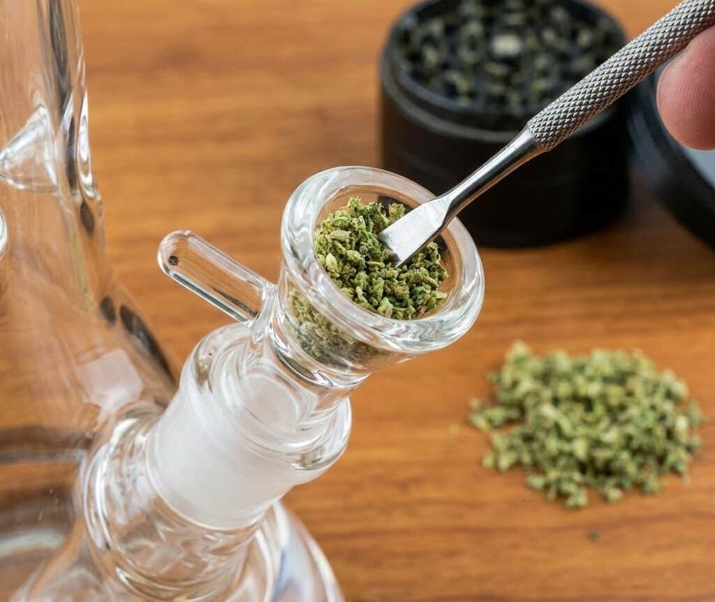 Close-up of a hand using a metal poker tool to carefully pack ground marijuana into a clear glass bong bowl on a wooden table.