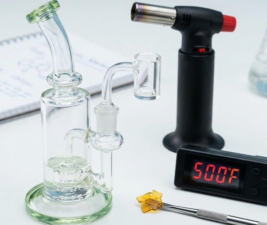 A glass dab rig, black butane torch, digital thermometer reading 500°F, and a dab tool with amber shatter on a white surface.