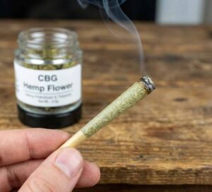 A close-up of a hand holding a lit hemp pre-roll with smoke rising from the tip, with a jar labeled "CBG Hemp Flower" resting on a wooden table in the blurred background.